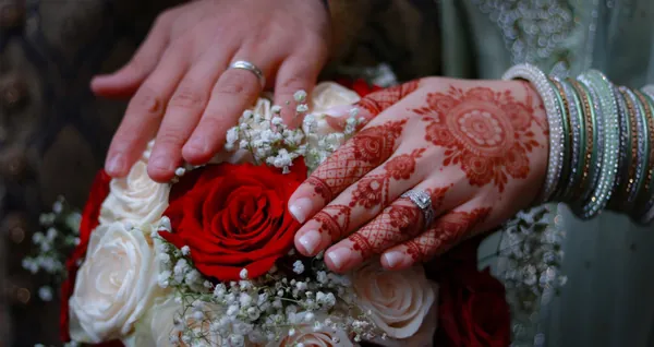 Bride’s hennaed hands resting on the groom’s hand over a red and white rose bouquet